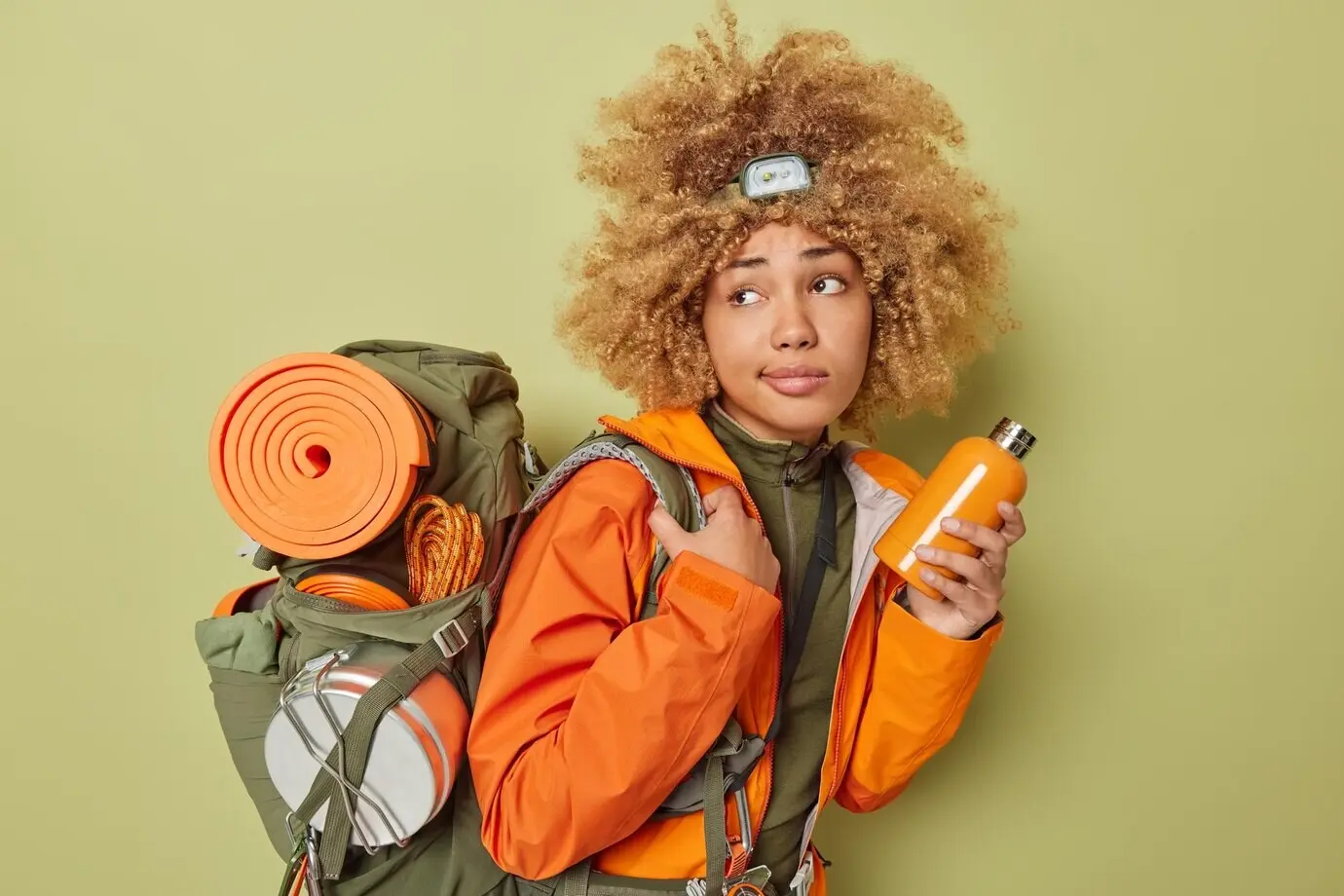 Seitliche Aufnahme einer nachdenklichen Camperin mit lockigem Haar, die eine Flasche mit frischem Wasser hält sowie eine orangefarbene Jacke und einen Rucksack trägt, posiert vor dunkelgrünem Hintergrund. Konzept: Wandern und Tourismus.