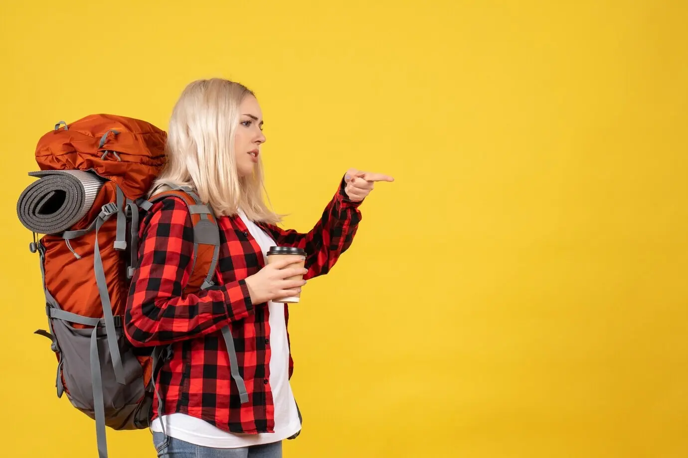 Frontansicht einer blonden Frau mit ihrem Rucksack, die einen Kaffeebecher hält und mit den Fingern nach rechts zeigt.